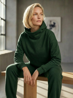 Model wearing Bou Clothing’s  Forest luxury Deryn hoodie and matching joggers, seated on a wooden bench in a softly lit modern interior, showcasing a relaxed fit, premium fabric, and minimalist neutral style.